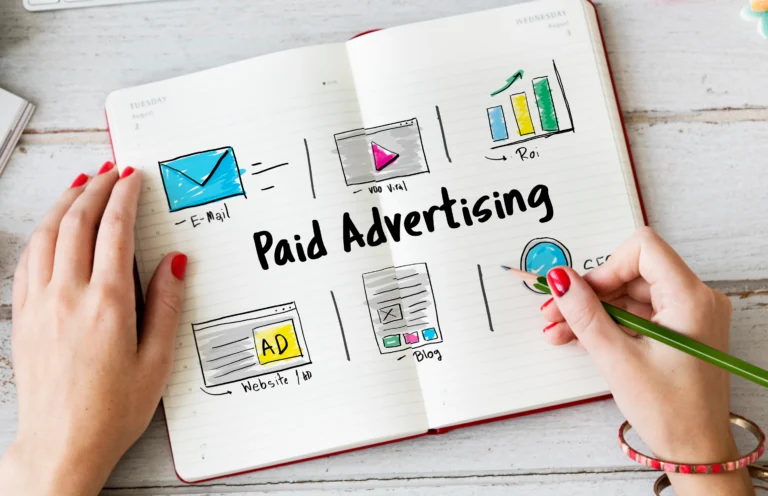 Top view of a notebook with hand-drawn illustrations labeled 'Paid Advertising,' including icons for email, video, SEO, website ads, and blog, with a person holding a pencil.