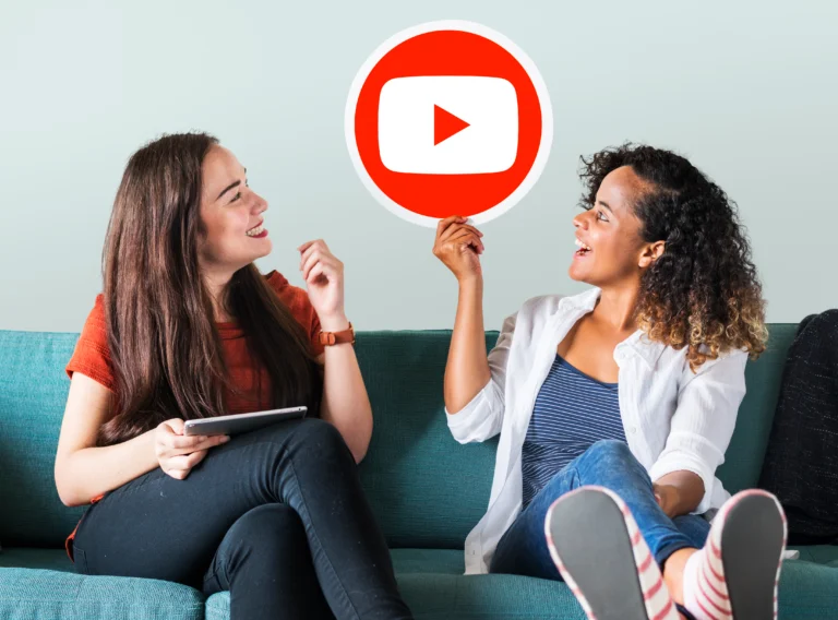 Two women sitting on a couch, smiling and discussing while holding a YouTube play button icon, representing YouTube Ads and video marketing strategy.