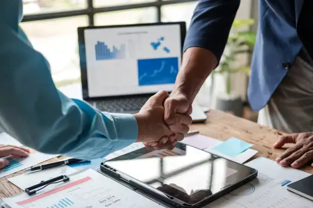 Two professionals shaking hands over a desk with laptops, charts, and marketing documents, symbolizing collaboration in WhatsApp and email marketing strategies.